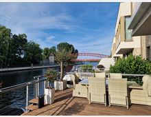 Terrasse mit Holzboden neben einem Fluss, ausgestattet mit hellen Rattanmöbeln und Pflanzen in Töpfen; im Hintergrund eine rote Brücke, dichtes Grün und moderne Wohngebäude.