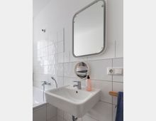 Weiß gefliestes Badezimmer mit einem Waschbecken, darüber ein rechteckiger Spiegel mit abgerundeten Ecken und ein kleiner runder Spiegel. Links befindet sich eine Badewanne mit Armaturen, und auf dem Waschbecken steht eine Flasche mit Flüssigseife. Ein Handtuch hängt an einem Handtuchhalter rechts.