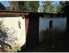 Kleines, weiß verputztes Gebäude mit brauner Holztür und kleinem rechteckigem Fenster; umgeben von Bäumen und Sträuchern. Eine Metalldose zum Füttern von Vögeln hängt an der Wand, und der Boden ist mit Laub bedeckt.