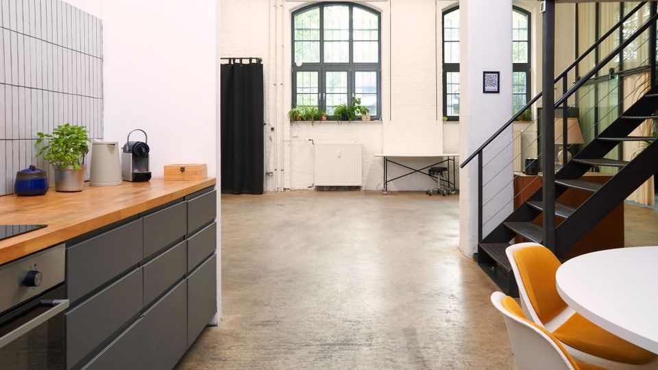 Heller, loftartiger Innenraum mit offener Küchenzeile links, Betonboden und weißen Wänden mit großen, bogenförmigen Sprossenfenstern. Rechts führt eine schwarze Metalltreppe an einer Glaswand vorbei; im Vordergrund steht ein runder Tisch mit Stühlen.