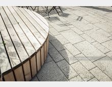 Runde Sitzbank aus hellem Holz auf einem gepflasterten Platz mit beigefarbenen Steinplatten; im Hintergrund sind Metallstühle und deren Schatten zu sehen.