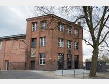 Backsteingebäude in einem industriellen Stil mit großen Fenstern und mehreren Etagen, umgeben von einem gepflasterten Vorplatz und einem Baum im Vordergrund. An der Gebäudefassade ist ein rundes Schild angebracht, und der Eingang ist über eine Rampe zugänglich.