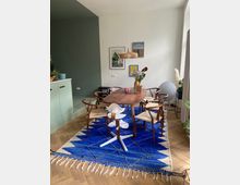 Esszimmer in modernem Stil mit einem rechteckigen Holztisch, umgeben von Stühlen aus Holz und Geflecht. Unter dem Tisch liegt ein blauer Teppich mit geometrischem Muster; im Hintergrund befinden sich eine grüne Wand, Dekorationen, Pflanzen und Bilderrahmen an der weißen Wand.