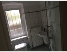 Kleines Badezimmer mit weißen Fliesen, einer Duschkabine mit Glaswand, einem Waschbecken und einer Toilette. Ein mattiertes Fenster lässt diffuses Licht herein, neben dem sich ein Heizkörper befindet.