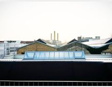 View over an urban rooftop with a long glass skylight on a metal roof in the foreground. Behind it are brick industrial buildings with sawtooth rooflines and distant smokestacks against a bright sky.