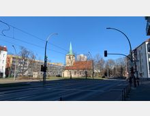 Straßenkreuzung mit Ampeln im Vordergrund, im Hintergrund eine kleine Backsteinkirche mit rotem Dach und grünem Kirchturm. Umgeben von Bäumen und moderner Architektur, unter klarem, sonnigem Himmel.