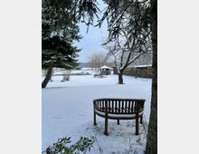 Schneebedeckter Garten mit einer hölzernen Rundbank im Vordergrund, umgeben von Bäumen und Sträuchern. Im Hintergrund ein kleiner Pavillon und gestapeltes Holz vor einer schneebedeckten Wiese.