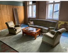 Wohnzimmer in einem teils renovierungsbedürftigen Altbau mit großen Sprossenfenstern, Holzvertäfelung an einer Wand und abblätterndem Putz. In der Mitte steht eine Sitzgruppe aus grünem Sofa und zwei Sesseln um einen niedrigen Holztisch auf einem großen Teppich.