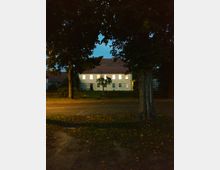 Ein zweistöckiges Gebäude mit heller Fassade und erleuchteten Fenstern, umgeben von Bäumen, aufgenommen bei Dämmerung. Im Vordergrund sind eine Straße und ein von Herbstlaub bedeckter Boden zu sehen.