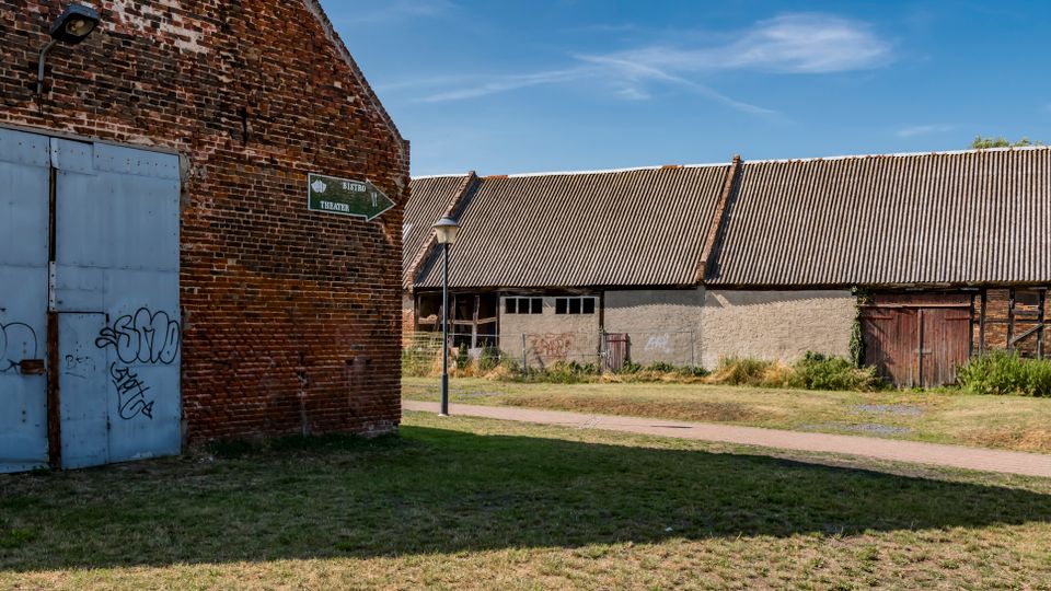 Hofbereich mit alten Wirtschaftsgebäuden: links eine Backsteinfassade mit großem grauem Metalltor und Graffiti, rechts ein langes Stall- oder Scheunengebäude mit Wellblechdach und Holztoren. Dazwischen verlaufen ein Grasstreifen und ein gepflasterter Weg mit einer kleinen Wegtafel und Laterne unter blauem Himmel.