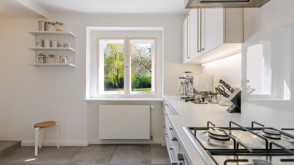 Helle, moderne Küche mit weißen Schränken, langer Arbeitsplatte und Gasherd unter einer Dunstabzugshaube. Ein großes Fenster über dem Heizkörper bietet Blick ins Grüne; links sind offene Wandregale und ein kleiner Hocker.
