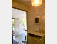 Heller Innenraum eines Wohnhauses mit Blick in einen angrenzenden Raum und weiter nach draußen auf eine Terrasse. Im Vordergrund steht ein kleines weißes Schrankmöbel mit Dekorflaschen, an der Decke hängt eine leuchtende Glaslampe.