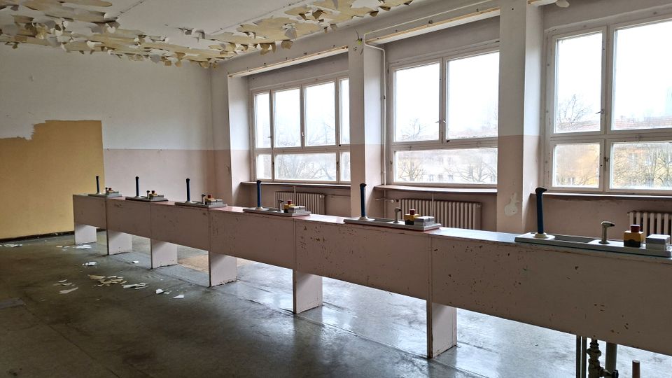 Verlassener, leerer Werkraum mit langen Reihen von Arbeitstischen und montierten Hebeln/Bedienelementen, vor großen Fenstern mit Heizkörpern darunter. Die Decke zeigt abblätternde Farbe, und der Betonboden ist stellenweise mit abgelösten Farbresten bedeckt.