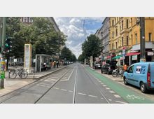 Straßenansicht mit Straßenbahngleisen in der Mitte, flankiert von Bäumen, Fußgängerwegen, einem Fahrradstreifen und Reihen von Wohn- und Geschäftsgebäuden im Altbaustil. Links steht eine Straßenbahnhaltestelle mit Wartehäuschen und Informationssäule, rechts parken Autos vor kleinen Läden und Cafés.