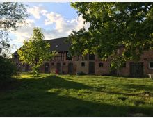 Langer Backsteinbau eines ehemaligen landwirtschaftlichen Hofes mit dunklem Satteldach und mehreren Holztoren und kleinen Fenstern. Davor liegt eine Wiese mit Bäumen, deren Äste den Blick teilweise einrahmen, unter einem Himmel mit vereinzelten Wolken.