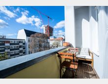 Ein schmaler Balkon mit Holzboden, ausgestattet mit einem kleinen Holztisch, klappbaren Stühlen und einer gepolsterten Sitzbank. Im Hintergrund sind mehrstöckige Wohnhäuser, ein großer Baum und ein Baustellengebäude mit roten Baukränen zu sehen, unter blauem Himmel mit wenigen Wolken.
