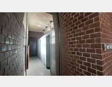 Enger Flur mit roten Ziegelwänden, einer Decke mit sichtbarer Abnutzung und weißen Türen auf der rechten Seite. Am Ende des Flurs befindet sich eine dunkle Tür.