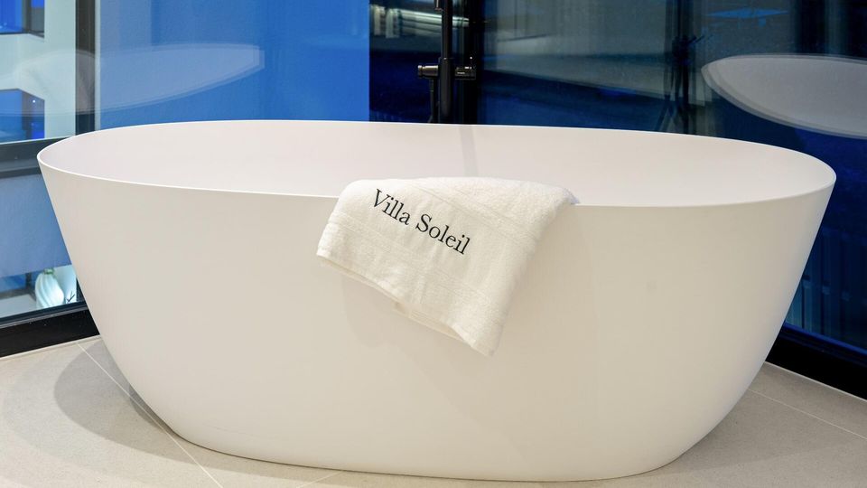 Moderne Badezimmeransicht mit einer freistehenden, ovalen weißen Badewanne auf hellen Fliesen. Dahinter sind eine schwarze Standarmatur und große Glasflächen mit blau getönten Spiegelungen zu sehen.