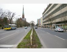 Straßenszene mit zwei Fahrspuren pro Richtung, getrennt durch einen Grünstreifen mit niedrigen Sträuchern und einer Metallbarriere. Links steht ein gelber Linienbus an einer Haltestelle, im Hintergrund ragt der spitze Turm einer Backsteinkirche empor, rechts flankiert ein modernes, mehrstöckiges Gebäude mit Geschäften die Straße.