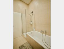 Modernes Badezimmer mit beigen Fliesen, einer weißen Badewanne mit Handbrause, einer verglasten Duschkabine, einer weißen Tür und einem beigen Teppich auf dem Boden.