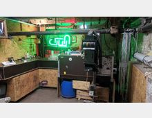 Ein rustikaler Raum mit rohen Holz- und Steinwänden, ausgestattet mit einer Bar, Flaschen, Neonbeleuchtung und einer veralteten Fotolaborvergrößerungsmaschine (Durst CLS 1000) im Vordergrund. Sichtbare Rohre und improvisierte Möbel verstärken den industriellen Kellercharakter.