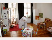 Ein helles Kinderzimmer mit Holzfußboden, einem kleinen Tisch mit bunten Stühlen, einem Papp-Spielhaus, einem Schaukelpferd und verschiedenen Spielzeugen. Der Raum hat große Fenster mit Vorhängen, darunter eine Fensterbank mit Pflanzen, sowie helle Möbel und eine Tür zu einem Balkon.