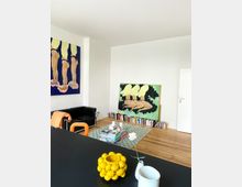 Wohnzimmer mit Holzboden, weißen Wänden und künstlerischem Flair. Zwei Gemälde in kräftigen Farben lehnen an den Wänden, ein schwarzes Ledersofa steht links, und ein niedriger Tisch mit Büchern befindet sich auf einem karierten Teppich; ein Regal aus gestapelten Büchern steht entlang der Wand.