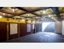 Unterführung einer U-Bahn-Station mit gelb gefliesten Wänden, dekorativen Ornamenten und einer Uhr über einer Infotafel. Rechts führt eine Treppe ins Tageslicht, links ist eine Reihe verriegelter Türen zu sehen.