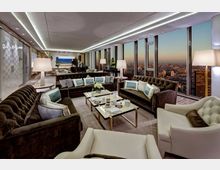 Luxuriös eingerichtetes Wohnzimmer in einem Hochhaus mit Panoramafenstern, die einen Ausblick auf eine Stadtlandschaft bei Sonnenuntergang bieten. Der Raum verfügt über elegante Sofas, Polstersessel, einen zentralen Couchtisch, Lampen und einen schwarzen Flügel im Hintergrund.