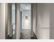 Schmaler, moderner Flur mit weißen Wänden, dunklem Holzfußboden und minimalistischem Design. Am Ende des Flurs hängt ein gerahmtes Kunstwerk an der Wand, links ist ein Geländer zu einem Treppenbereich sichtbar.