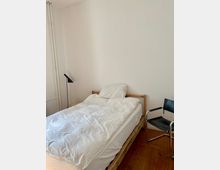 Schlichtes Schlafzimmer mit einem einfachen Holzbett, weißer Bettwäsche, einer schwarzen Stehlampe und einem schwarzen Stuhl mit Metallrahmen. Der Raum hat Holzboden und weiße Wände.