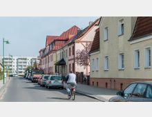 Eine Wohnstraße mit Reihenhäusern in pastellfarbenen Fassaden und roten Ziegeldächern auf der rechten Seite. Autos parken entlang der Straße, während ein Radfahrer auf der Fahrbahn fährt, und moderne Wohngebäude sind im Hintergrund sichtbar.