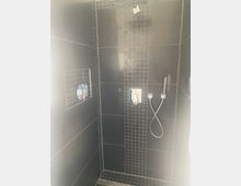 Geflieste, bodengleiche Dusche mit dunklen Wandfliesen und einem vertikalen Streifen aus kleinen Mosaikfliesen in der Mitte. Sichtbar sind eine Regendusche an der Decke, eine Handbrause mit Schlauch sowie eine eingelassene Wandnische mit Ablage.