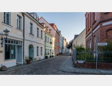 Schmale Kopfsteinpflastergasse mit historischen Gebäuden, darunter farbige Wohnhäuser mit dekorativen Fassaden auf der linken Seite und ein rotes Backsteingebäude rechts. Ein Straßenschild mit der Aufschrift „Steingasse“ ist sichtbar, umgeben von einer niedrigen Metallumzäunung und Grünpflanzen.