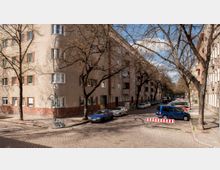 Straßenszene in einem Wohnviertel mit mehrstöckigen Wohnhäusern aus Putz und Backstein im unteren Bereich. Am Straßenrand stehen geparkte Autos unter kahlen Bäumen, und eine temporäre Straßenabsperrung ist im Vordergrund sichtbar.