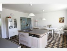 Helle Küche mit Schachbrettboden, einer Kücheninsel mit Spülbecken im Vordergrund und einem langen Esstisch mit Stühlen im Hintergrund. An der weißen Wand hängen minimalistische Lampen, daneben steht ein weißer Schrank, und eine türkisfarbene Holztür verleiht dem Raum einen rustikalen Akzent.