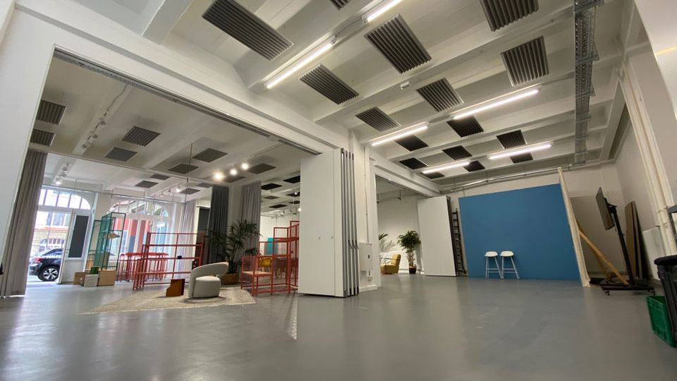 Großes, helles Studio- oder Veranstaltungsloft mit hoher Decke, sichtbaren Trägern, Akustikpaneelen und langen Deckenleuchten über glattem grauem Boden. Im Raum stehen mobile Trennwände, ein blauer Hintergrund, Sitzmöbel und Pflanzen; große Fensterfronten geben den Blick nach draußen frei.