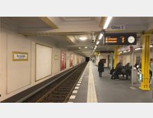 U-Bahn-Station mit Gleis und Bahnsteig in Berlin, beschildert als „Kochstraße – Checkpoint Charlie“. Die Wände sind weiß mit gelben Rahmen und Werbeplakaten, gelbe Stützpfeiler und eine digitale Anzeige der Abfahrtszeiten sind sichtbar, einige Personen warten auf Sitzbänken.