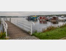 Eine hölzerne Fußgängerbrücke mit Geländer führt zu einem Steg, der sich über einen ruhigen See erstreckt. Im Hintergrund befinden sich bunte Hausboote und Yachten, umgeben von bewaldetem Ufer und grauem Himmel.