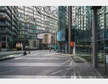 Moderne urbane Passage mit Hochhausfassaden aus Glas und Stahl, teilweise reflektierend, und einem großen digitalen Display. Im Vordergrund eine gepflasterte, offene Fläche mit einer Person, die auf einer Bank sitzt, umgeben von Glasstrukturen und Eingängen zu Geschäftsräumen.