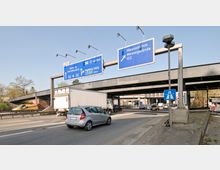 Straßenansicht einer mehrspurigen Kreuzung in Berlin mit Autobahnschildern, die auf Ziele wie Hamburg, Charlottenburg, Magdeburg und das Messegelände (ICC) hinweisen. Im Hintergrund ist eine aufgeständerte Brücke sichtbar, während Autos und Lkw die Straße befahren.