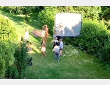 Eine Wiese in einem Garten mit mehreren Menschen, die an einer Fotoshootingszene arbeiten. Im Hintergrund befindet sich eine große weiße Leinwand, umgeben von dichten, grünen Büschen und Bäumen.