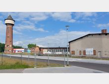 Backstein-Wasserturm neben einem Industrie- und Lagerkomplex mit verschiedenen Gebäuden, einer Zufahrtsstraße und Absperrzäunen im Vordergrund. Im Hintergrund ein moderner Gewerbebau und Grünflächen unter einem bewölkten Himmel.