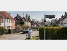 Straßenszene in einem Wohngebiet mit Einfamilienhäusern und Vorgärten. Links und rechts der gepflasterten Straße stehen farbige Häuser mit roten Ziegeldächern, während Autos am Straßenrand parken; im Vordergrund ein Straßenschild mit der Aufschrift „Flötnerweg“.