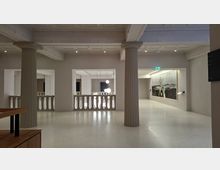 Heller Museums- oder Galerieinnenraum mit hoher Kassettendecke, breiten Unterzügen und zwei großen, geriffelten Säulen. Offene Balustraden zum angrenzenden Bereich, moderne Hängelampen und ein großformatiges Wandbild an der rechten Wand.