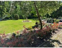 Ein gepflegter Garten mit einer großen Rasenfläche, einer kleinen Steinfigur auf einem runden Brunnen in der Mitte und blühenden Sträuchern in einem Blumenbeet im Vordergrund. Im Hintergrund stehen hohe Bäume, die den Garten umrahmen.