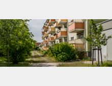 Mehrstöckiges Wohngebäude mit mehreren Balkonen in Orange- und Beigetönen, umgeben von Grünflächen, Sträuchern und Bäumen. Rechts neben den Gebäuden führt ein schmaler Weg durch die Gartenanlage.