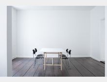Minimalistisch eingerichteter Raum mit weiß gestrichenen Wänden und dunklem Holzboden, in dessen Mitte ein weißer Tisch mit Holzgestell und vier schwarzen Stühlen steht. Im Hintergrund befindet sich eine helle Wand ohne Dekoration.