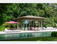 Moderner Pavillon mit großen Glasfronten inmitten eines Gartens, umgeben von dichtem Grün. Im Vordergrund ein Pool, daneben Sitzgelegenheiten mit weißen Polstern und rosa Sonnenschirmen.