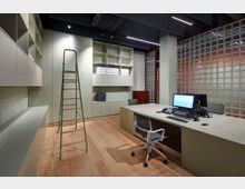 Modernes Büro mit einem großen Schreibtisch, einem Computermonitor und Büromaterialien. Die Rückwand besteht aus hohen Einbauschränken und Regalen mit Aktenordnern, daneben ein gesicherter Tresor; rechts ist eine Wand aus Glasbausteinen zu sehen, auf dem Holzfußboden steht eine Leiter.